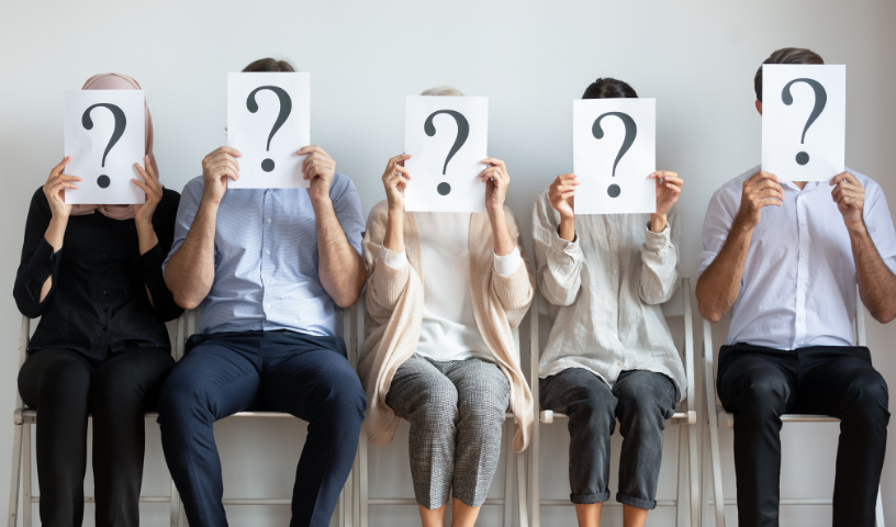 Line of people sat down, facing camera, holding a piece of paper with a question mark over their face