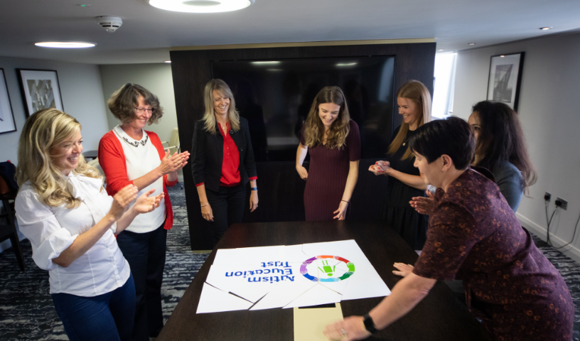 7 member of the Autism Education Trust Team, stood around a puzzle of the logo clapping, pieces of the puzzle pushed together.