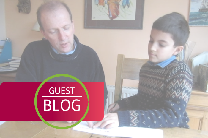 Guest blog symbol on top of faded photo of parent and child sat at kitchen table reading book together