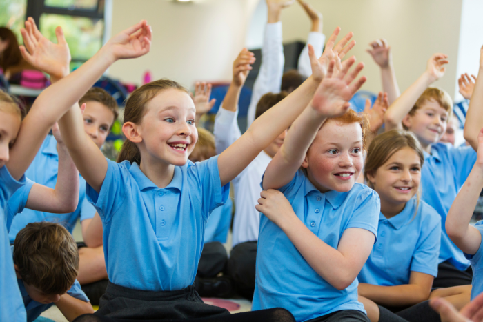Primary school pupils sitting in an assembly, smiling with their hands in the air.