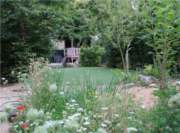 An outdoor playground with wooden climbing frame. The playground has lots of green space and some flowers blooming.