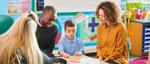 Parents sit on either side of their child in a classroom, facing a practitioner. They are all looking down at a picture in front of the child.