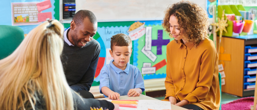 Parents sit on either side of their child in a classroom, facing a practitioner. They are all looking down at a picture in front of the child. 