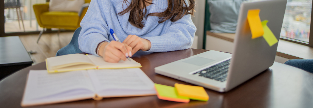 Close up of someone writing at their desk. Their laptop is also in front of them.