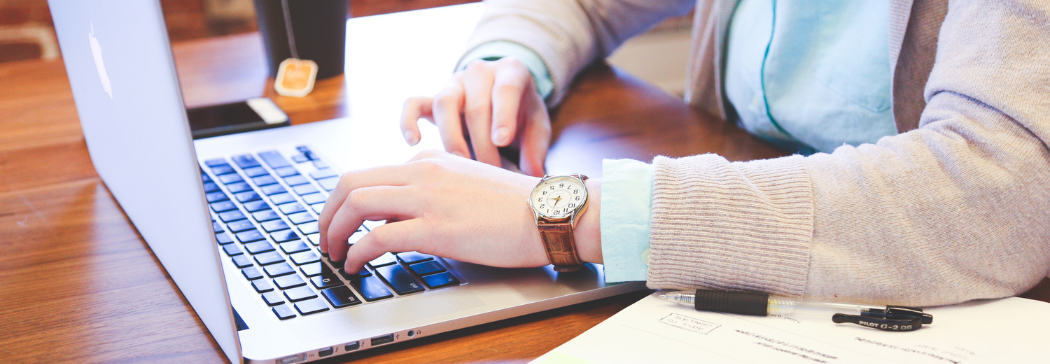 Close up of someone sitting at their desk, their hands are resting on their laptop keyboard. Their notebook and phone sits on either side of the laptop.