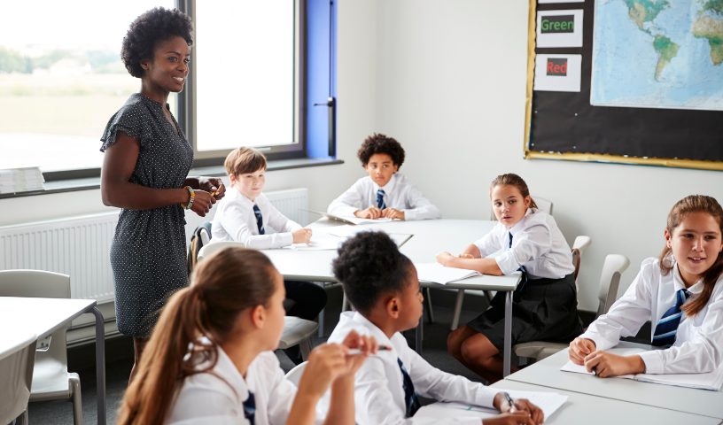 A teacher stands in a classroom with secondary school pupils sitting at their desks. The pupils are in white shirts and blue striped ties.