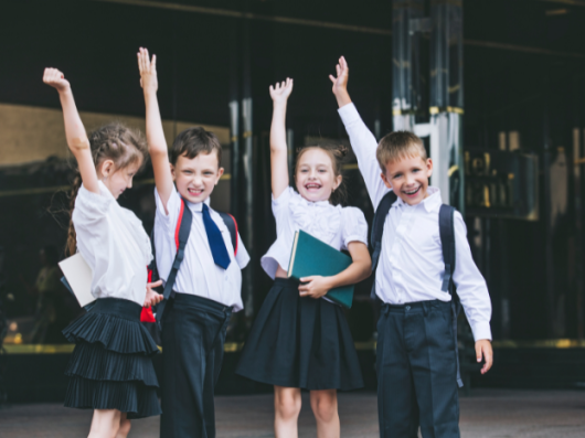 Four primary school pupils stand next to eachother outside school. They are all holing one hand in the air, some of them are smiling.