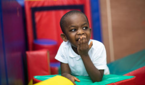 Child leaning on soft play, hand over mouth and looking around