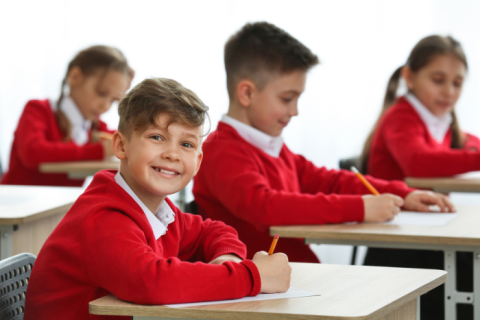 Pupils in exam room in red school uniform. One looking across room at camera, smiling.