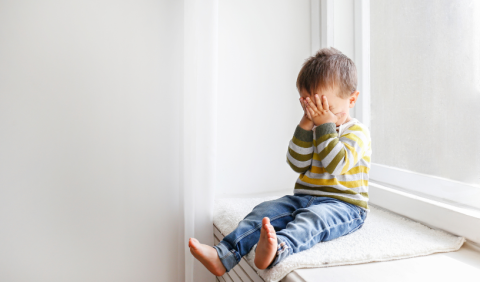 A toddler sits on a windowsill with their hands held over their face.