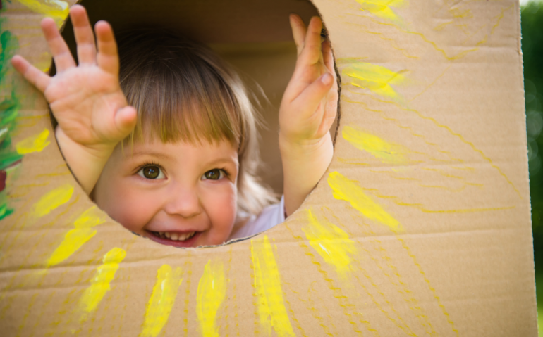 Toddler, smiling, peeking out of circle cut in a cardboard box, painted as a sunshine