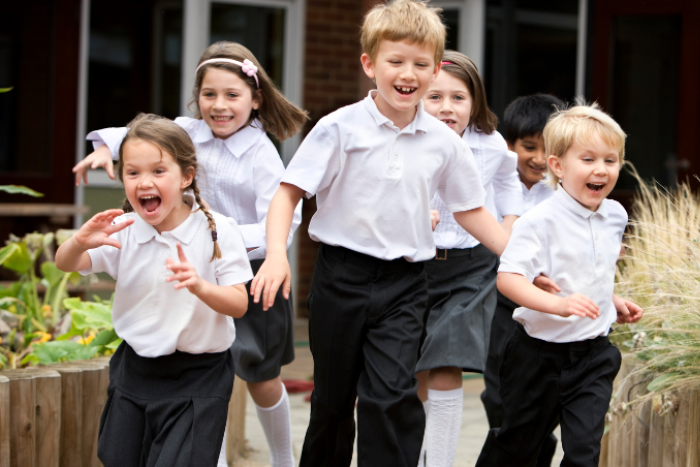 A group of primary school pupils in white polo shirts are running through a playground. They are all smiling and some of them cheering.
