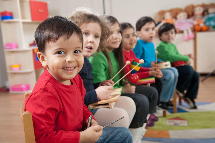 A group of early years children sit in a row. They are all holding percussion sticks. The child at the end of the row is looking at the camera and smiling.
