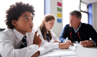 Boy in classroom looking ahead thoughtfully, teacher and classmate in background
