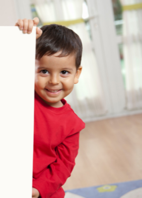 Child peering around white board at camera