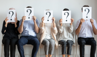 Line of people sat down, facing camera, holding a piece of paper with a question mark over their face