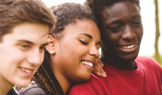 Three college teenagers, standing close, looking at something together, smiling.