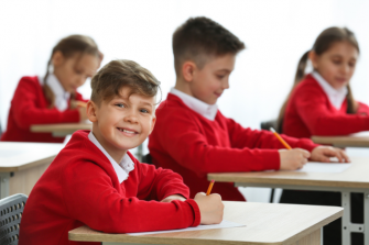 Pupils in exam room in red school uniform. One looking across room at camera, smiling.
