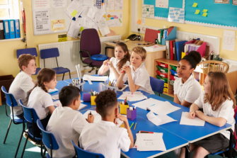 Eight primary school aged students sitting around table in classroom, chatting, paper and coloured pencils in front on them.