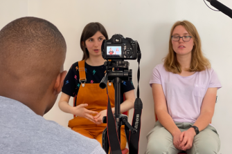 Two people sitting in chairs in front of camera, person behind the camera filming