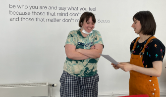 Two people standing up, talking whilst looking at piece of paper, smiling and chatting
