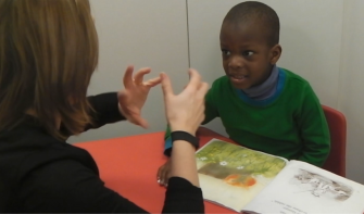 Young pupil sitting with teacher, talking to each other with book on the table between them