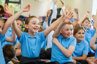 Primary school pupils sitting in an assembly, smiling with their hands in the air.