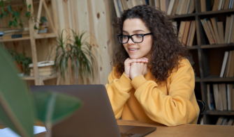 Young professional sitting at their desk at home, chin in hands, looking at laptop, smiling.