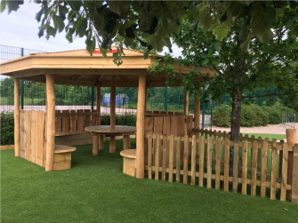 A wooden canopy with wooden benched inside. The canopy is on green astroturf.