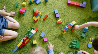 A group of children sit around colourful playing blocks. Some of them reach into the middle to pick one up. 