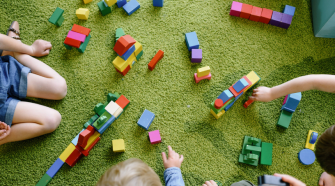 A group of children sit around colourful playing blocks. Some of them reach into the middle to pick one up.