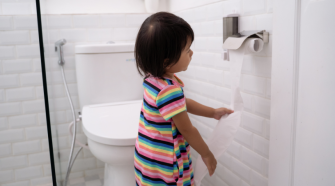 A toddler stands in front of the toilet and pulls on the end of the toilet roll.