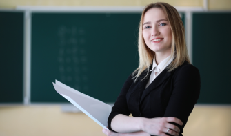 A teacher holding a handful of paper looks at the camera and smiles.