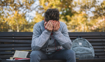 A young person sitting on a bench with their backpack and book sitting next to them. They have their head in their hands.