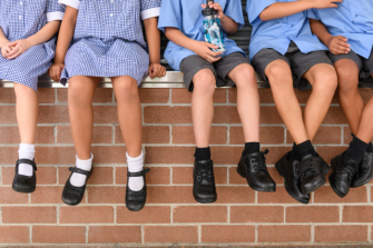 A group of primary school pupils sit on a wall in a row. Their legs dangle over the edge and you can see they shiny leather school shoes.