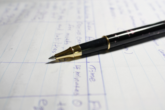 Pen resting on piece of paper, blurred text behind it