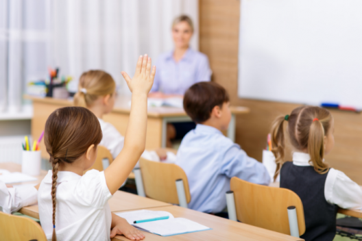 Primary school children in uniform sitting in classroom at desks, facing teacher. One pupil with hand raised.