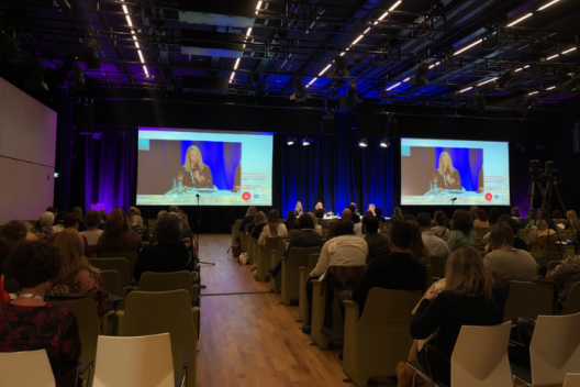 Sarah Broadhurst, Autism Education Trust Director, on screen at the front of conference room at Autism Europe, delegates in chairs looking at screen. 