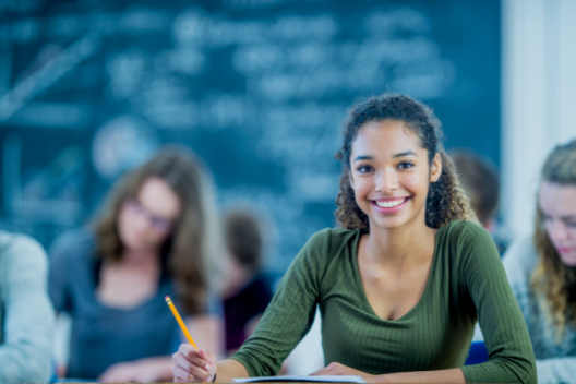 Pupil in exam hall, sitting at a table with exam paper in front of them, looks up at the camera, smiling. 