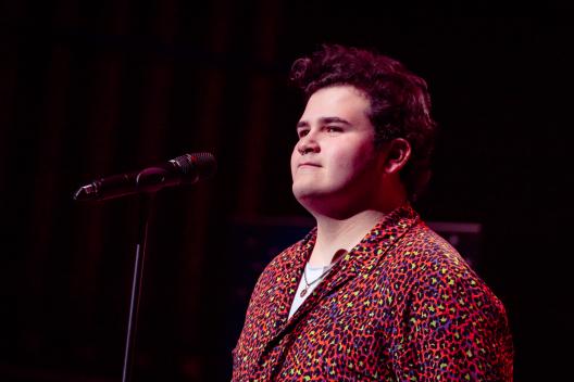 Performer standing on stage in front of a microphone. They are smiling. 