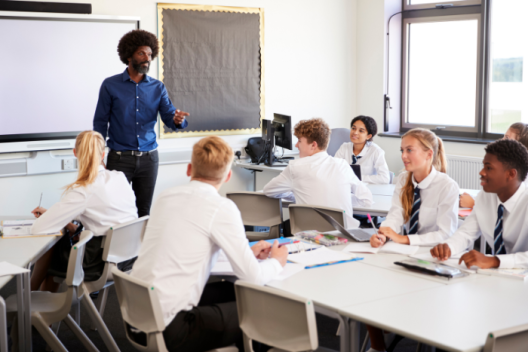 Teacher standing at the front of a classroom in front of pupils in school uniform. The pupils are smiling. 