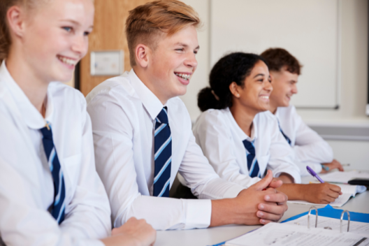 Group of young people in white shirt and blue striped tie. They are sitting at their desks in a classroom, smiling and laughing. 