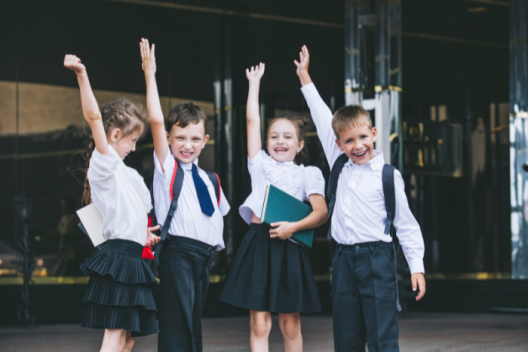 Four primary school pupils stand next to eachother outside school. They are all holing one hand in the air, some of them are smiling.