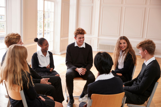 A group of students sit on chairs in a circle. They are looking at each other, talking and smiling. 