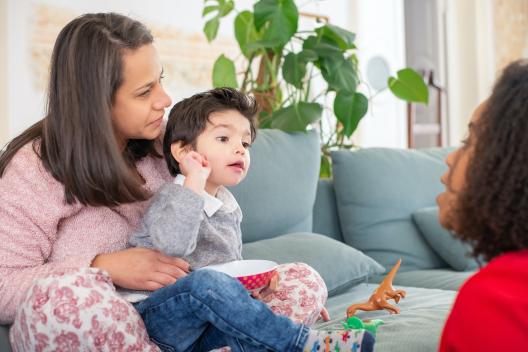 Early years child sits in adults lap. They are looking ahead of them, at another child.