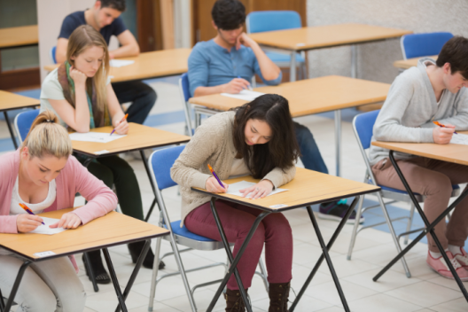 Students in an exam hall. They are all hunched over their individual tables, reading and writing on their exam papers.