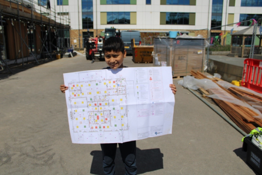 A young person stands on a building site, holding a large blue print