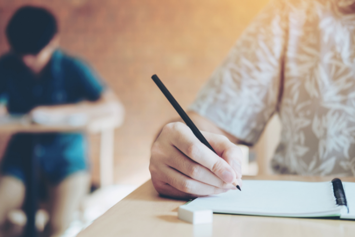 Close up on pencil and notepad, child in background concentrating, looking down at own desk.