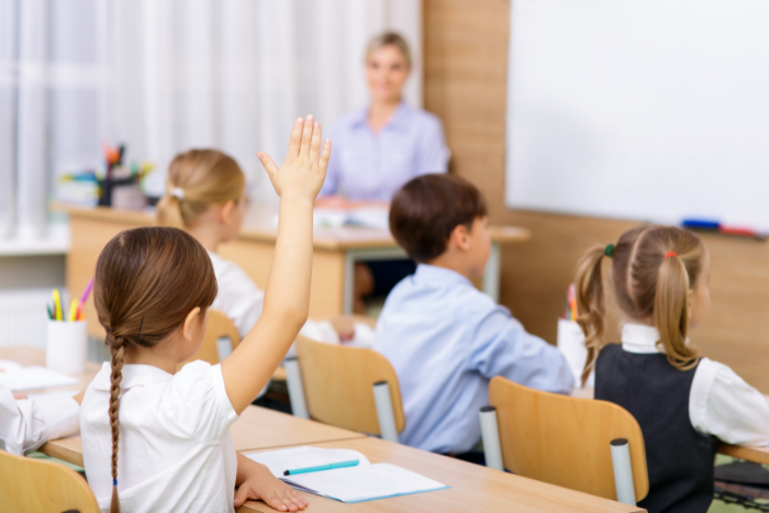 Primary school children in uniform sitting in classroom at desks, facing teacher. One pupil with hand raised.