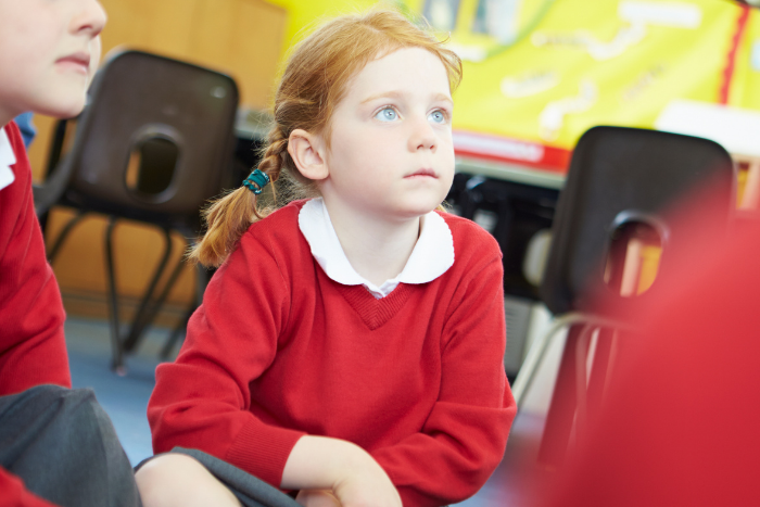Primary school pupil in white shirt and red jumper, sitting cross legged in classroom, looking up